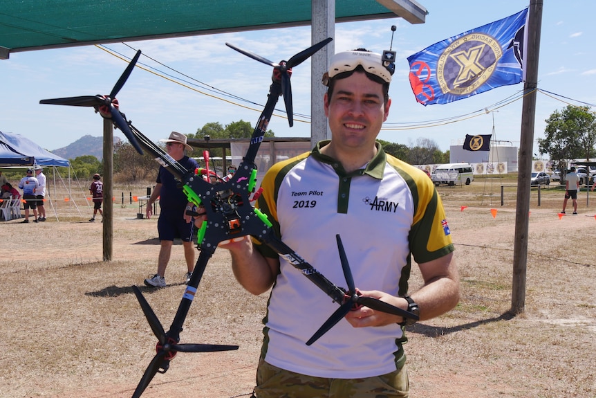 A man in a white and yellow t-shirt holds a large black and green drone, smiling with goggles on his head