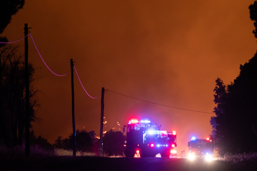 Two fire trucks lit against a fiery orange sky with power lines nearby