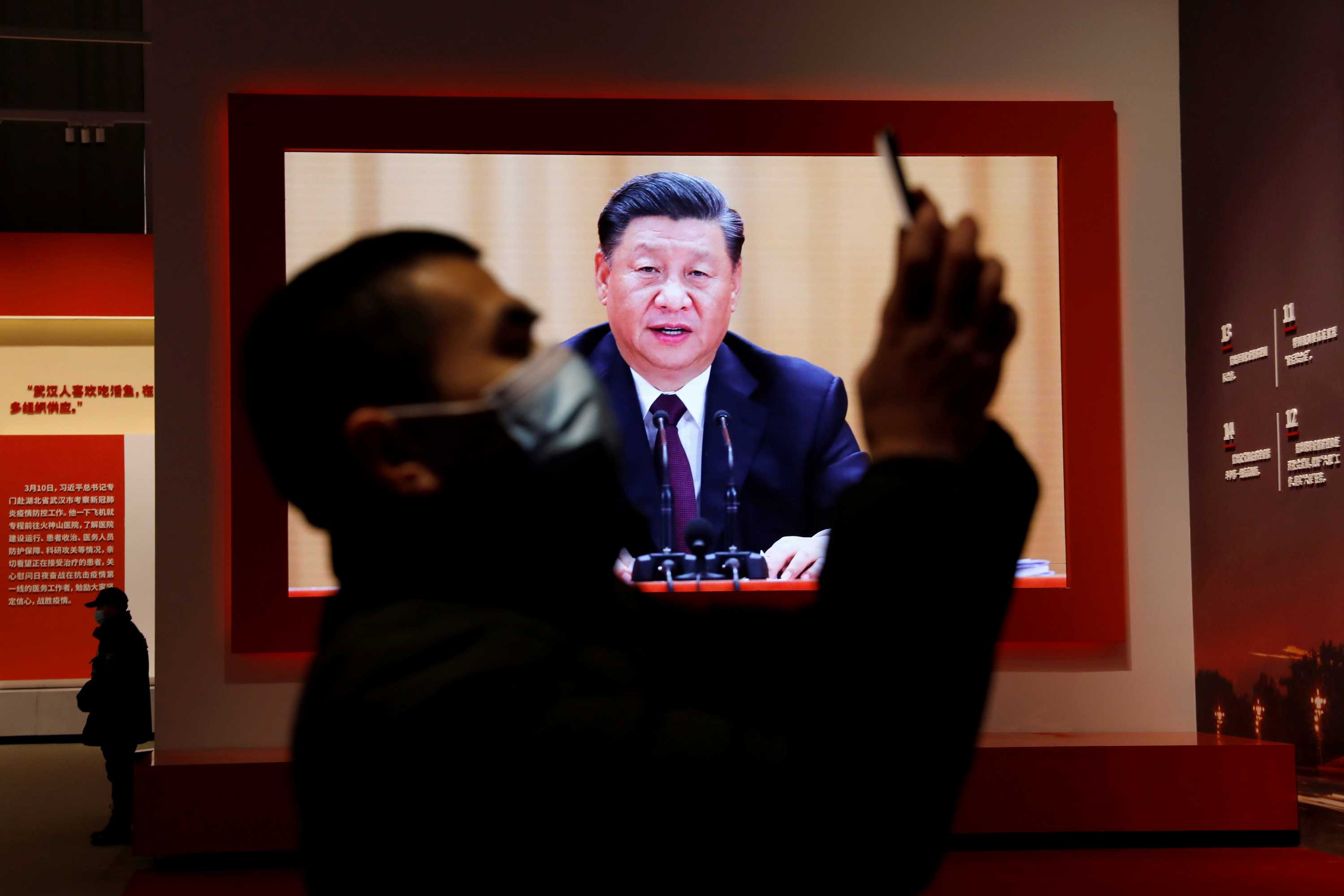 一名戴着口罩的男子在电视前手持手机，正在播放中国中年男子的讲话。