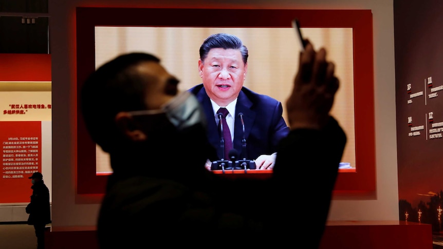 A man wearing a mask holds a mobile phone in front of TV showing speech of middle-aged Chinese man.