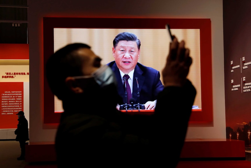 A man wearing a mask holds a mobile phone in front of TV showing speech of middle-aged Chinese man.