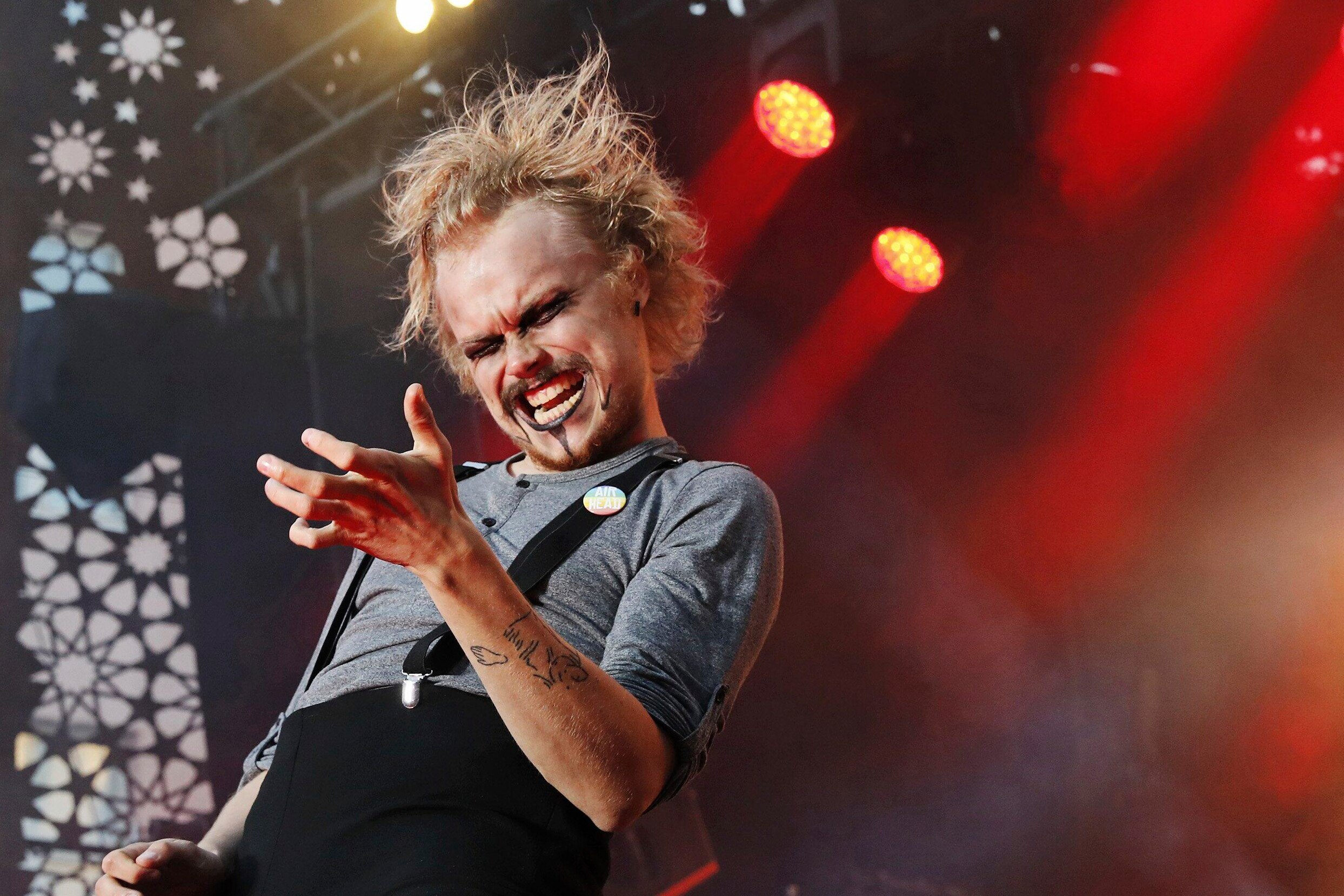 Close up of a man in black overalls and heavy makeup playing air guitar on a stage with red lights behind him