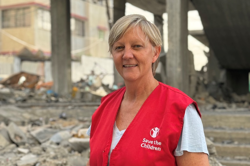 A woman with short blonde hair, wearing a red vest with a 'Save the Children' logo on it. She is standing in front of rubble.