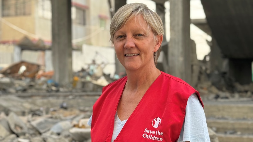 A woman with short blonde hair, wearing a red vest with a 'Save the Children' logo on it. She is standing in front of rubble.