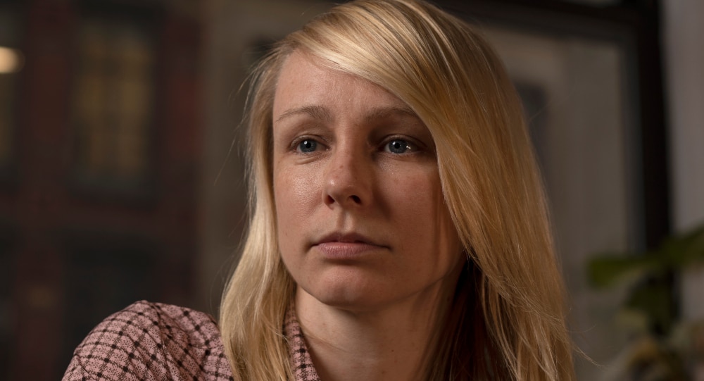 A woman with long blonde hair wears pink check button up shirt looks to the left and sits near apartment window in the evening.