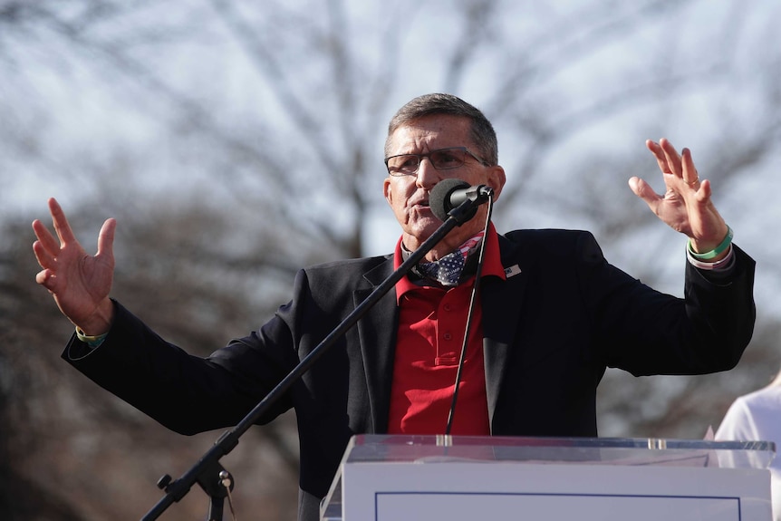 A man wearing a black jacket and red shirt holds his arms out as he speaks into a microphone.