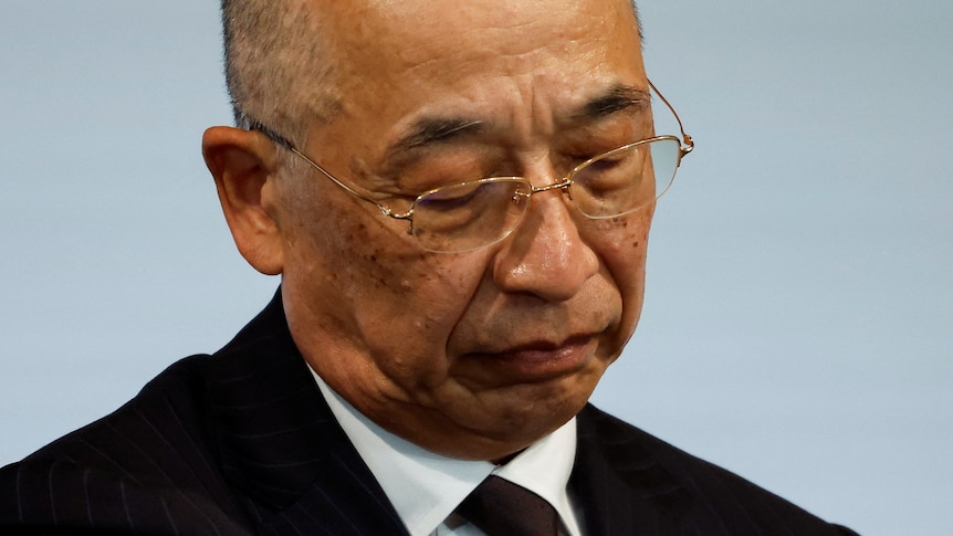 A close-up photo of an older Japanese man wearing a suit and glasses and looking forlorn.