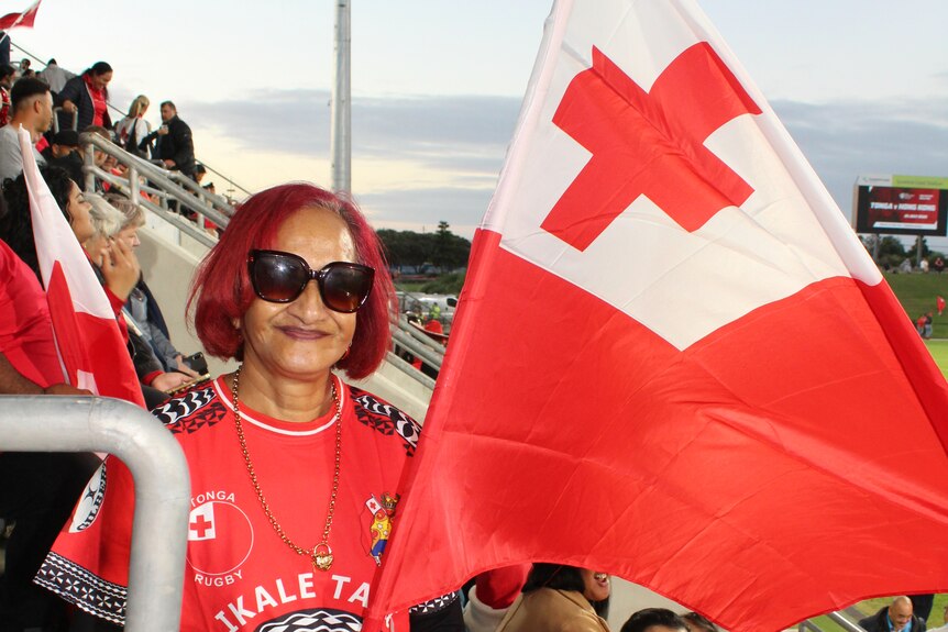 Une femme portant des lunettes de soleil tient un drapeau tongien dans les gradins d'un stade.