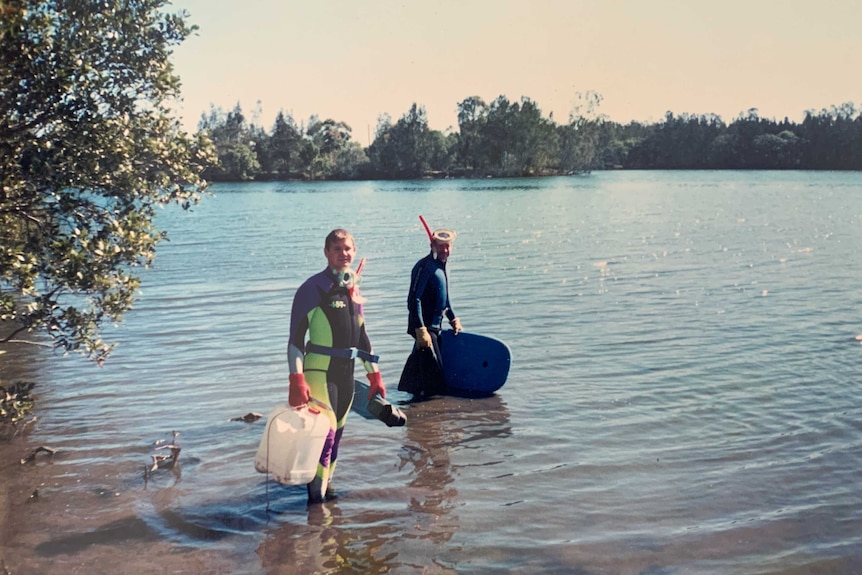 Two men standing in shallow water near a riverbank, wearing wetsuits, masks and snorkels.