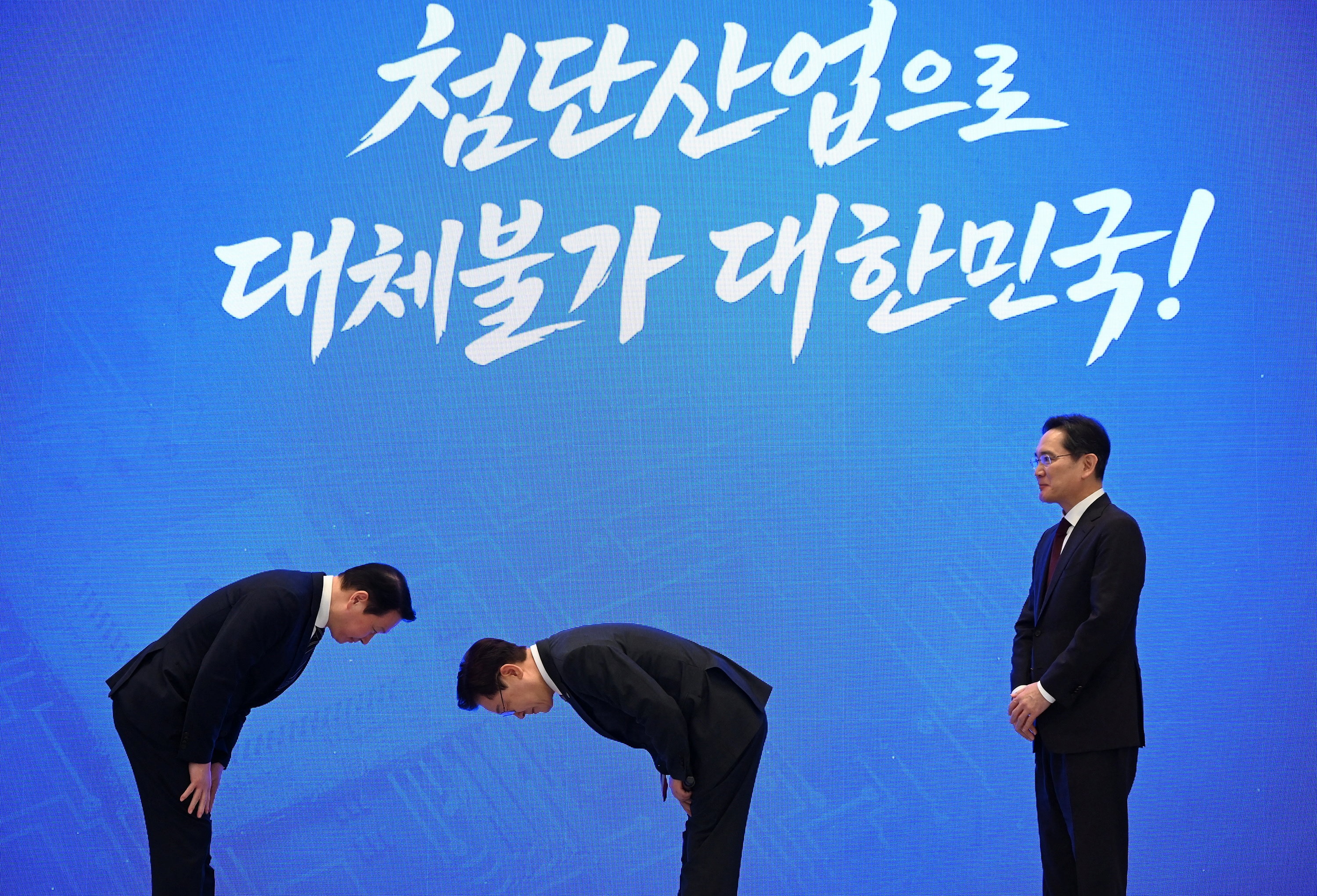 Two suited men bow deeply to each other on a stage as a third suited man watches on.