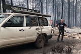 A man stands by a car with a sign that reads "climate action now or...?" in front of a burnt-out house.