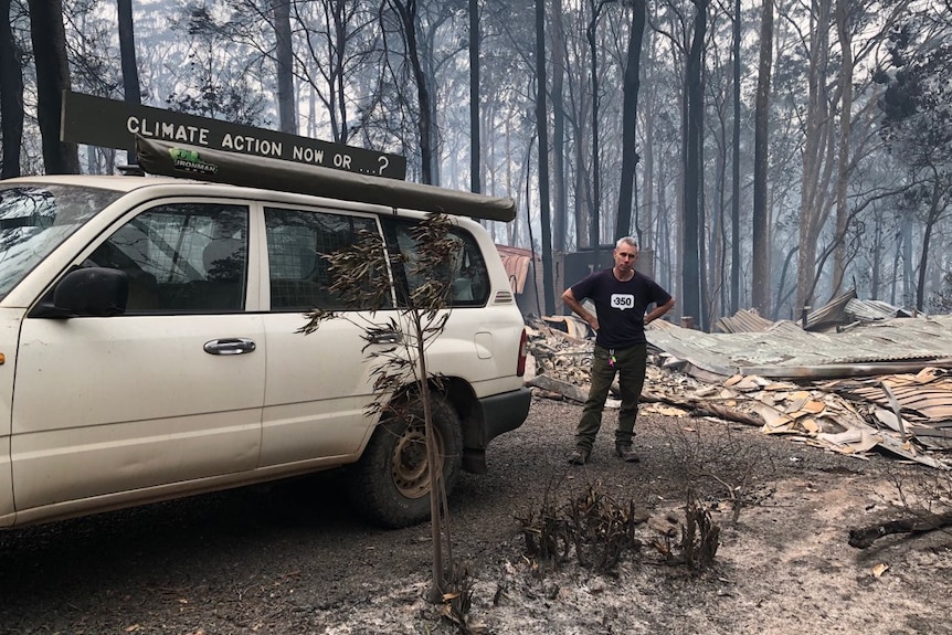 A man stands by a car with a sign that reads "climate action now or...?" in front of a burnt-out house.