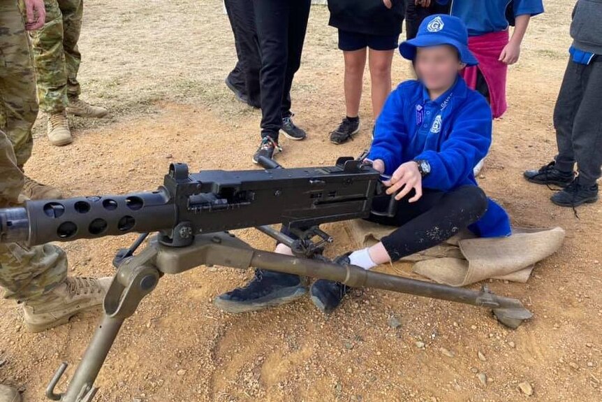 A child in school uniform sits behind a large weapon mounted on legs. They are overseen by two soldiers