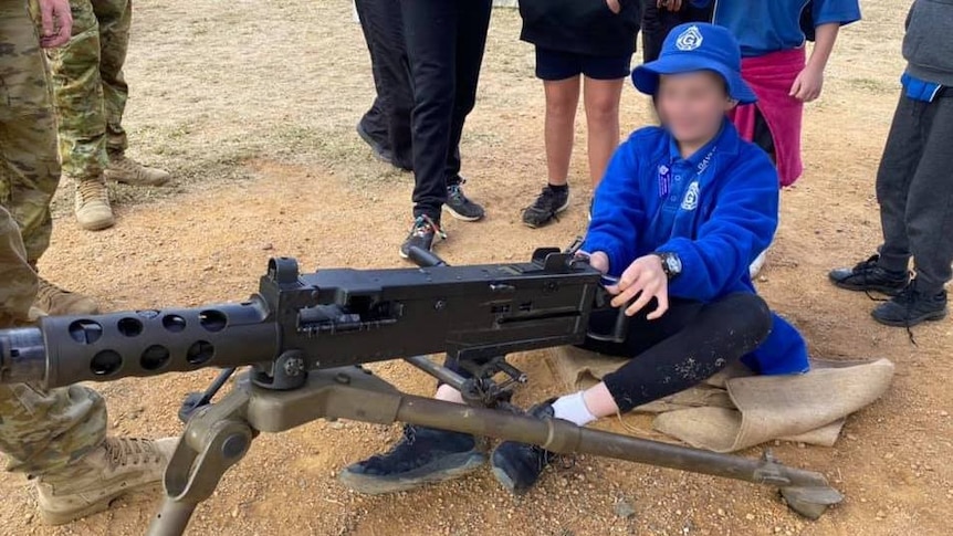A child in school uniform sits behind a large weapon mounted on legs. They are overseen by two soldiers