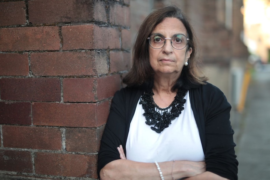 A woman wearing glasses, leaning against a brick wall, looks up to the side.
