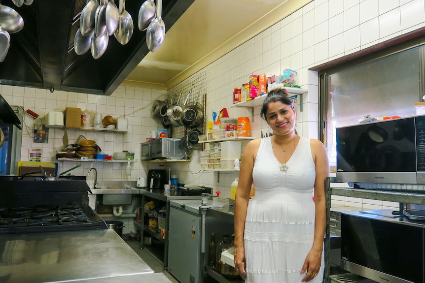 Geetu stands in a white dress in the motel's industrial kitchen, she is surrounded by ingredients and utensils.