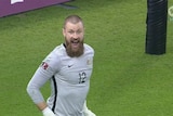 A bearded goalkeeper in a grey shirt stands with hands on hips, his eyes wide and mouth wide open after a penalty shootout.