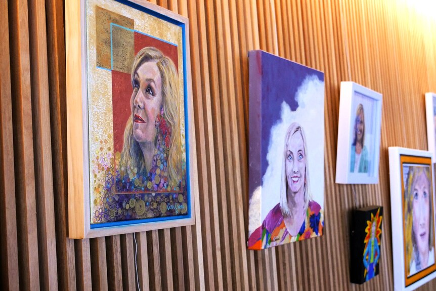 Colourful paintings of a woman with blonde hair are hang up in a jumbled line on a wooden wall.