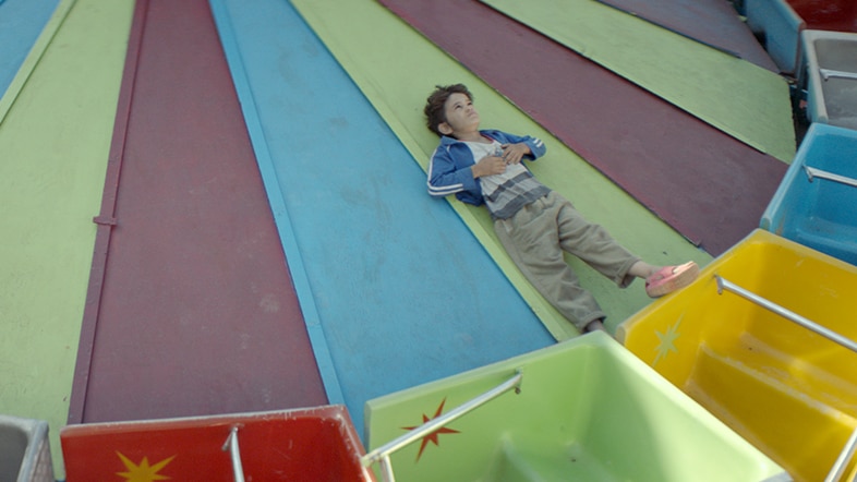 Colour still of Zain Al Rafeea lying on colourful amusement park ride in 2018 film Capharnaüm.