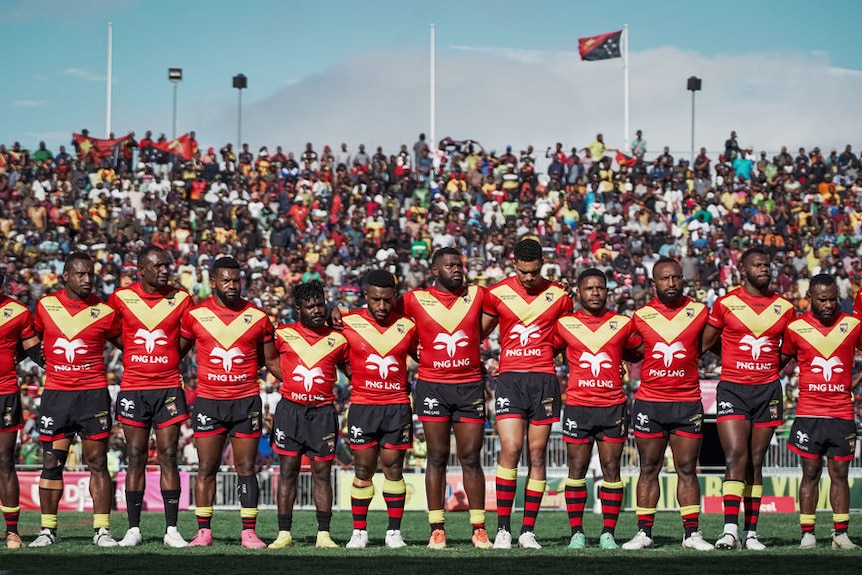 巴布亚新几内亚足球场上，一群身穿红黄黑球衣的足球运动员手挽手站立