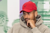Nick Kyrgios looks smug at a Wimbledon press conference, while wearing a red cap and a hoodie.
