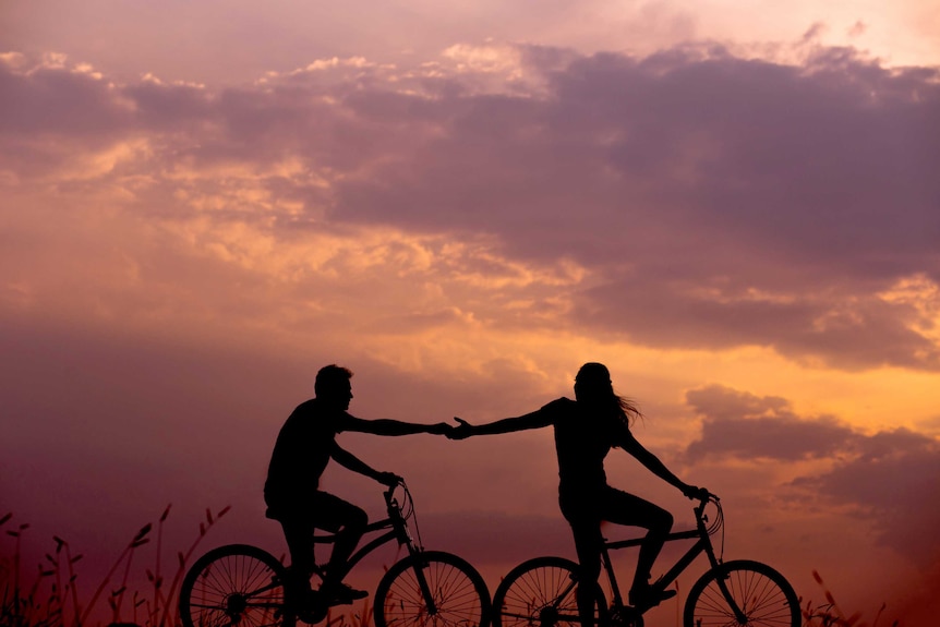 The silhouette of a young couple of high school sweethearts cycling through a field in front of the sunset.