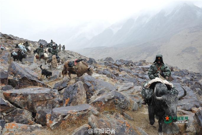 Across a foggy mountainous landscape, you view a group of Chinese military officers riding yaks traversing boulders.