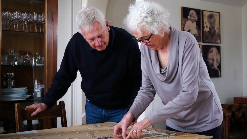 An older man and women with gray hair leaning over a table and placing a piece into a jigsaw puzzle.