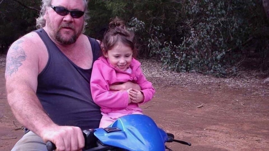 Stephen in a tank top and jeans riding a quad bike, his daughter Jess on his knee.