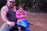 Stephen in a tank top and jeans riding a quad bike, his daughter Jess on his knee.