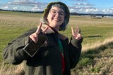 Is Hay is standing in a country field. They are doing a peace sign with both hands and smiling.