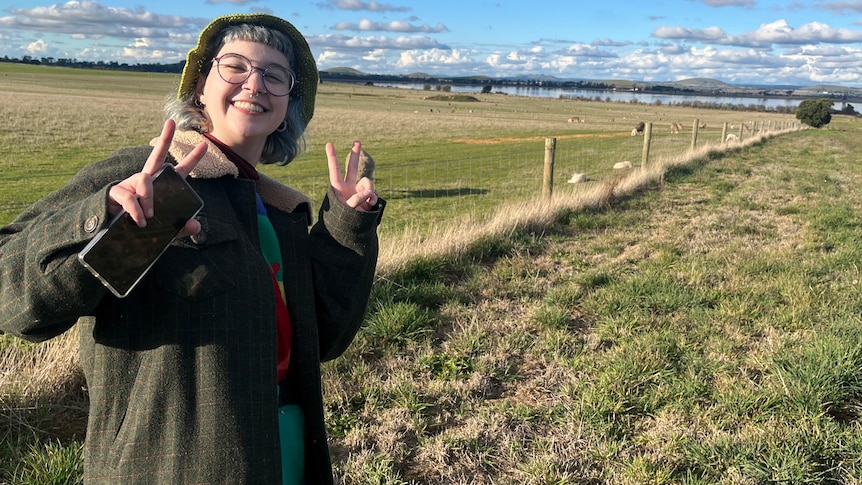 Is Hay is standing in a country field. They are doing a peace sign with both hands and smiling.