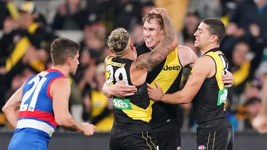 A star AFL forward is hugged by teammates after kicking a goal during a game. 