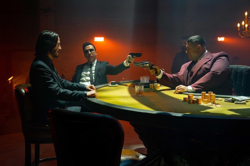A white man in a black suit, an Asian man in a black suit and a white man in a maroon suit hold guns around a poker table.