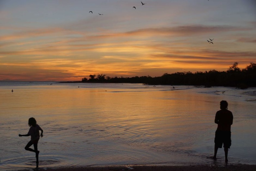 People stand in the shallows on Croker Island while the sun sets.