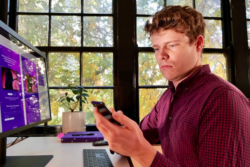 Hew Sandison looks at a phone while sitting in front a computer monitor