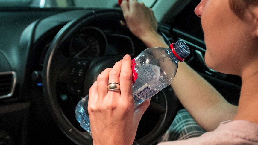 A tight interior shot of a woman drinking water at the wheel of a car.