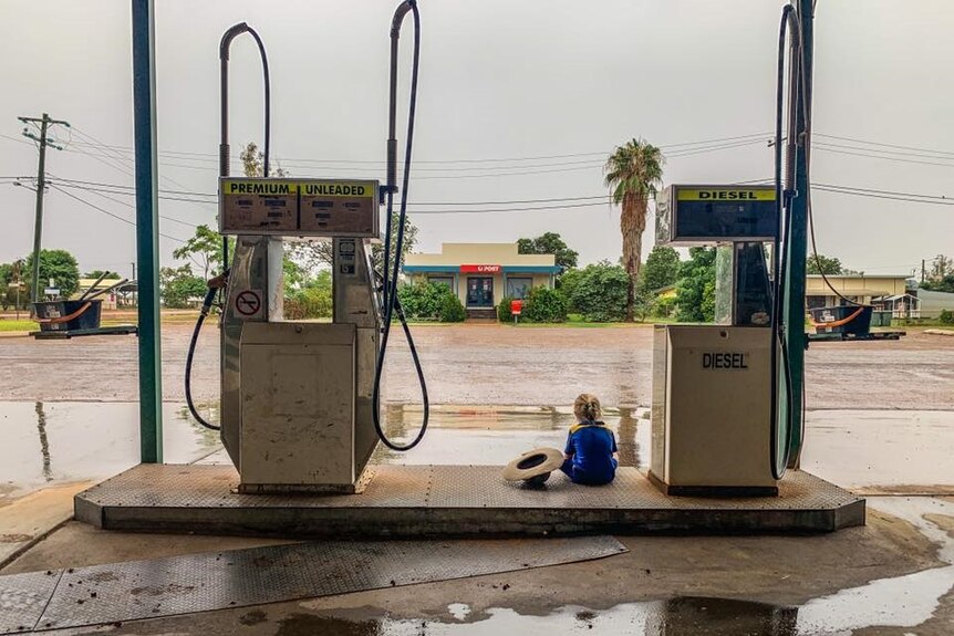 Child sits between two petrol bowsers at Windorah Service Station watching floodwaters.