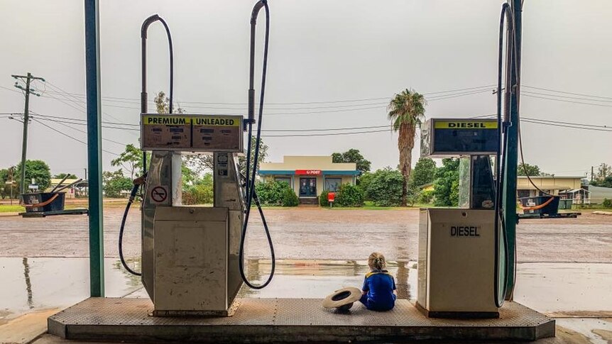 Child sits between two petrol bowsers at Windorah Service Station watching floodwaters.