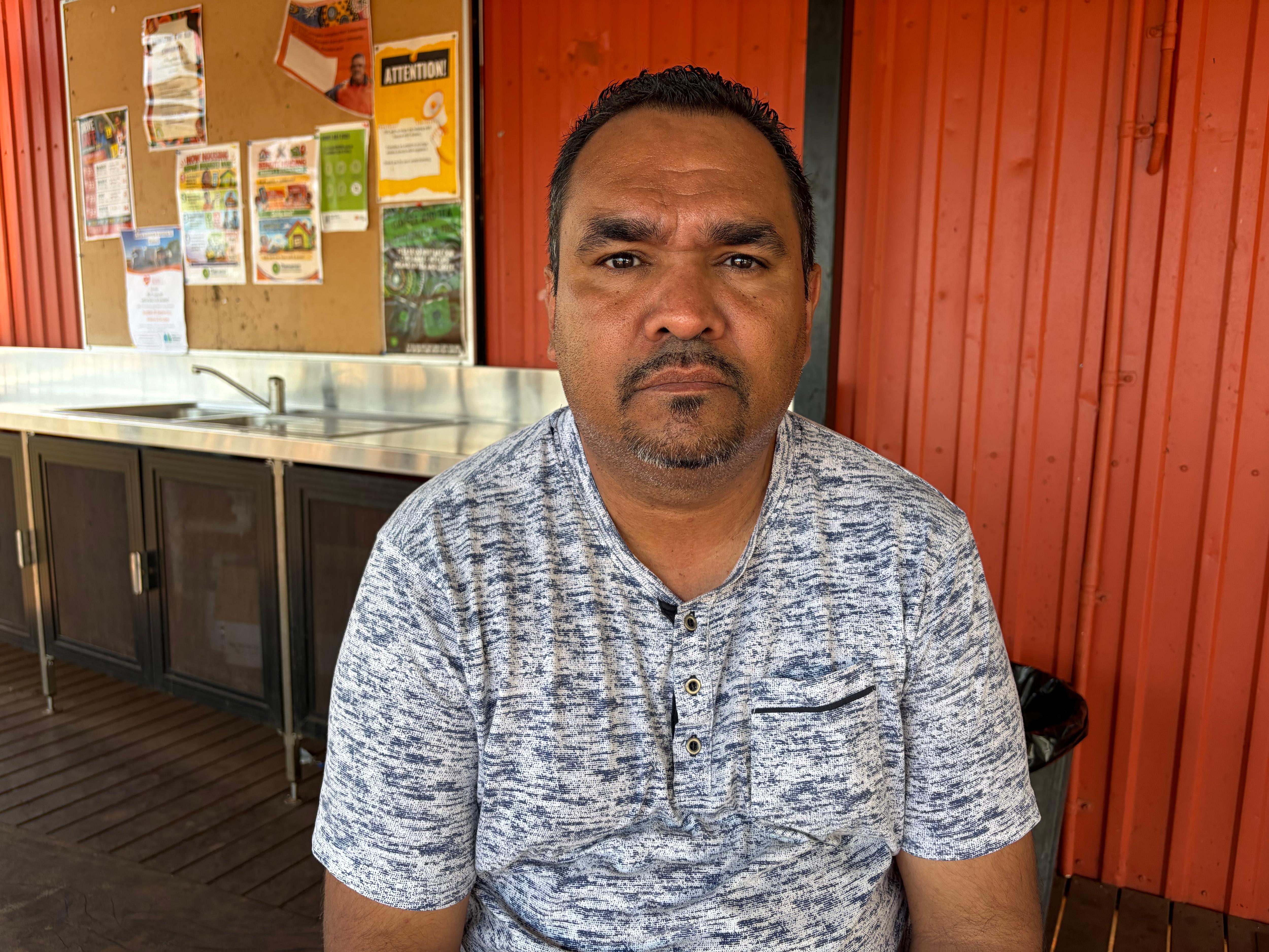 An Indigenous man, Mark Tunmuck-Smith, sits in front of red shed and noticeboard.