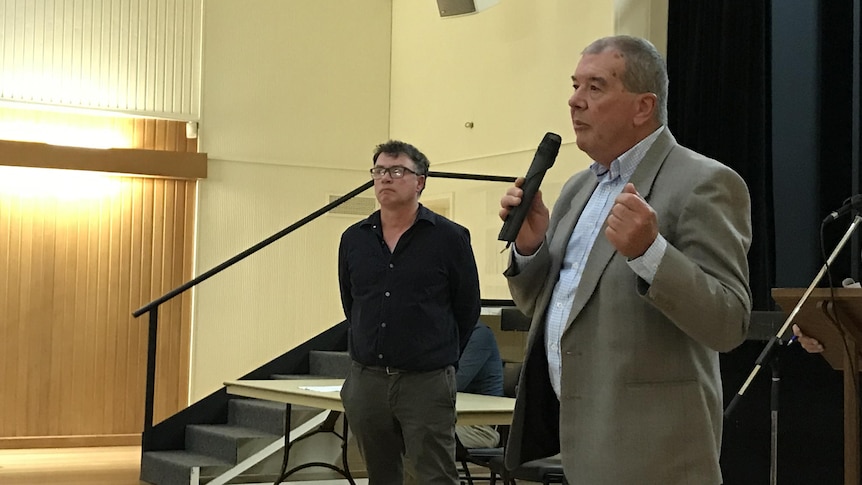 A man in a light-coloured sports coast speaks into a microphone in a municipal hall.