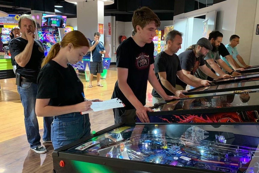 A row of men playing pinball machines and a young woman standing looking at a clipboard in front of the machines.