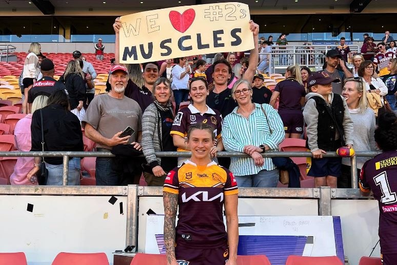 Brisbane Broncos player Julia Robinson stands in front of fans holding up a sign reading "We love #2's muscles"