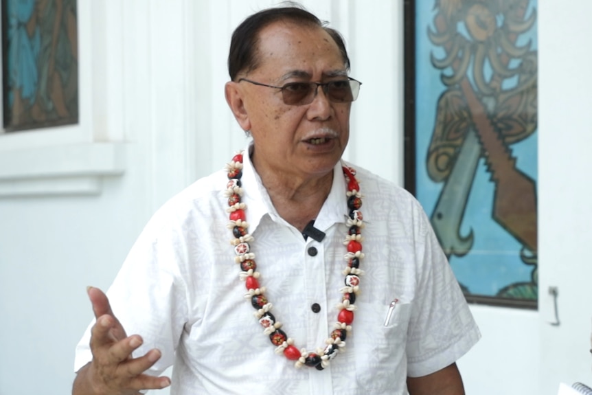 Salausa Dr John Ah Ching in a white shirt and red, white and brown necklace looks off-camera during an interview.