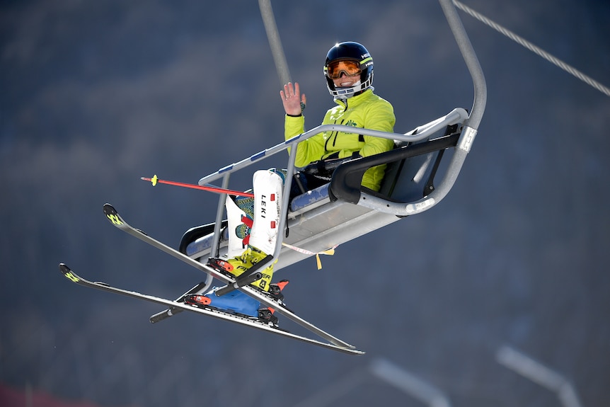 Rae Anderson is sitting on a chair lift, she smiles and waves at the camera.