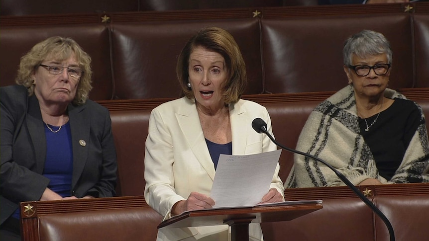 Nancy Pelosi stands at a lectern speaking with two people sitting behind her.