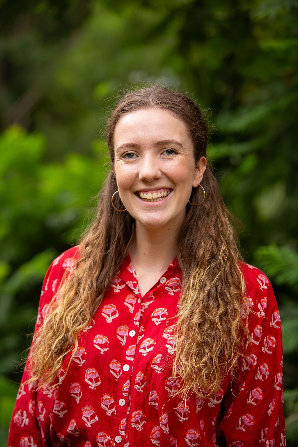 A smiling woman with wavy red  hair stands in front of some greenery.