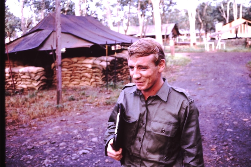 Man in uniform with a clipboard and military base in background.