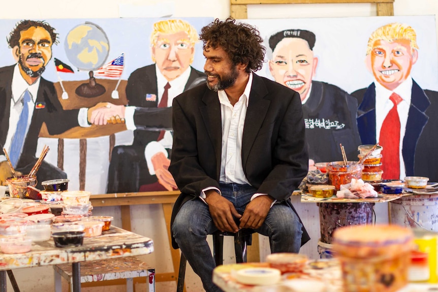 A man sits in the middle of a room with paintings behind him