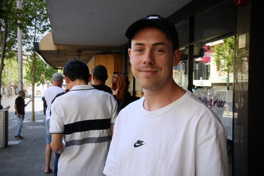 A man in a white t-shirt stands in a queue.